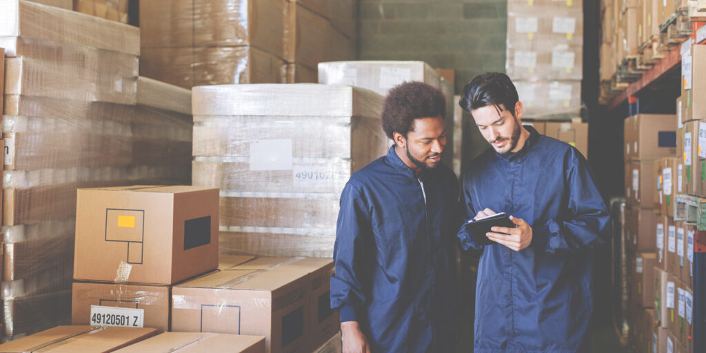 Two warehouse workers reviewing a tablet among stacked boxes