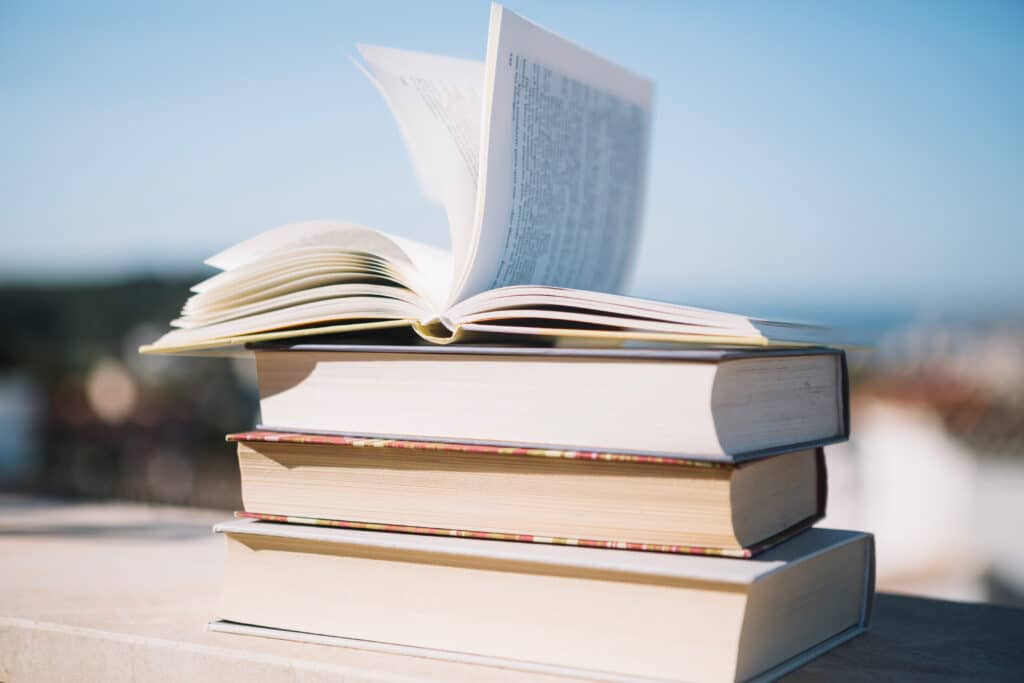 Open book with pages fluttering in the wind, stacked on two closed books outdoors under a blue sky.